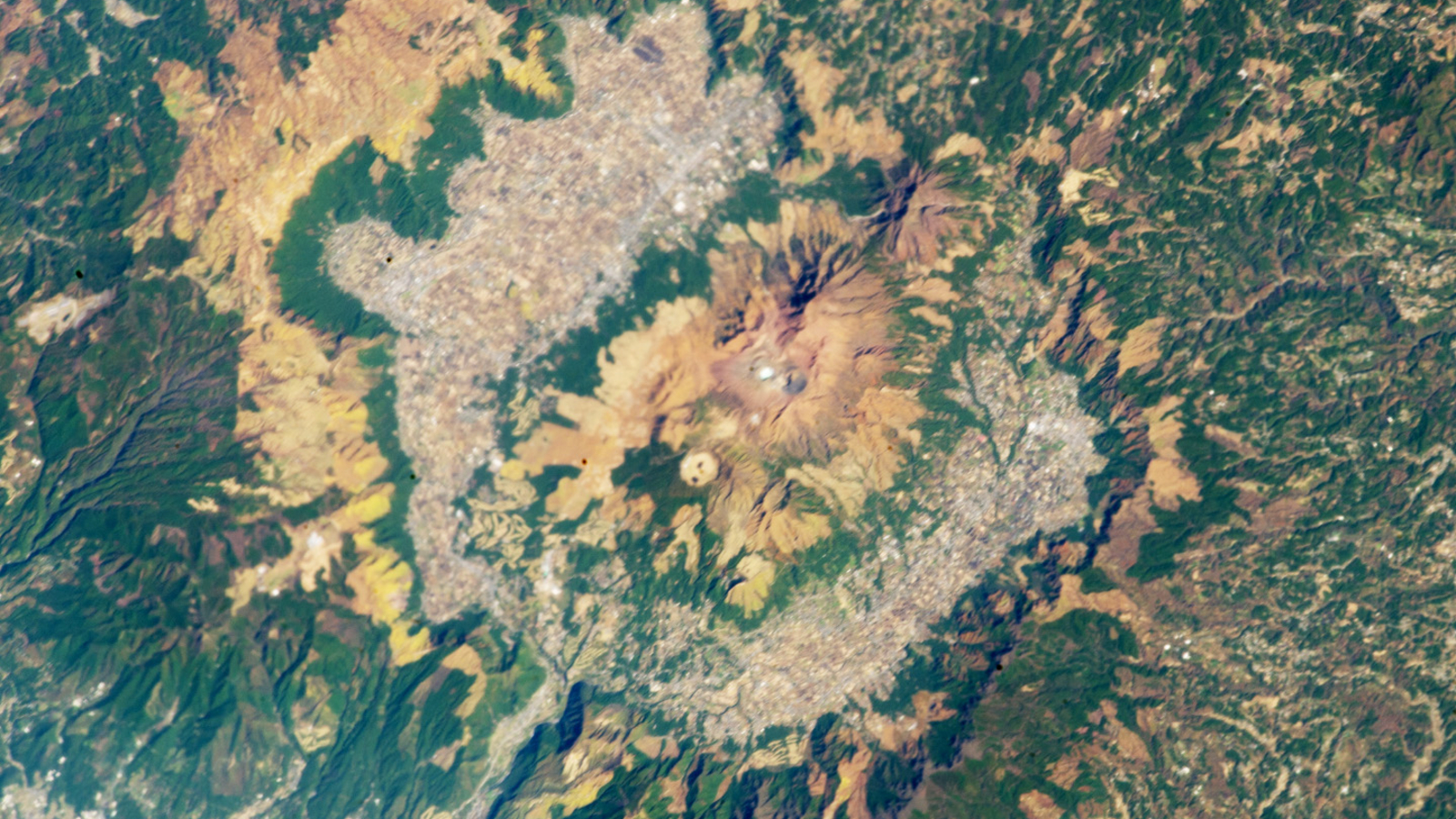 An astronaut photo of a giant caldera in Japan. A central cluster of volcanoes is surrounded by a ring of urban and agricultural land and a larger ring of ridges